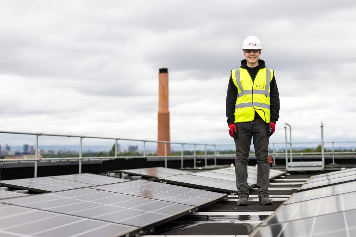 A employee stood on a roof next to a solar panel wearing a helmet and highvis